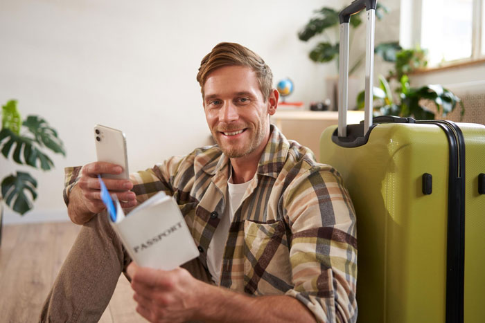 Man holding passport and phone, smiling next to suitcase, reflecting on teens preferring ex over dad for Europe trip planning