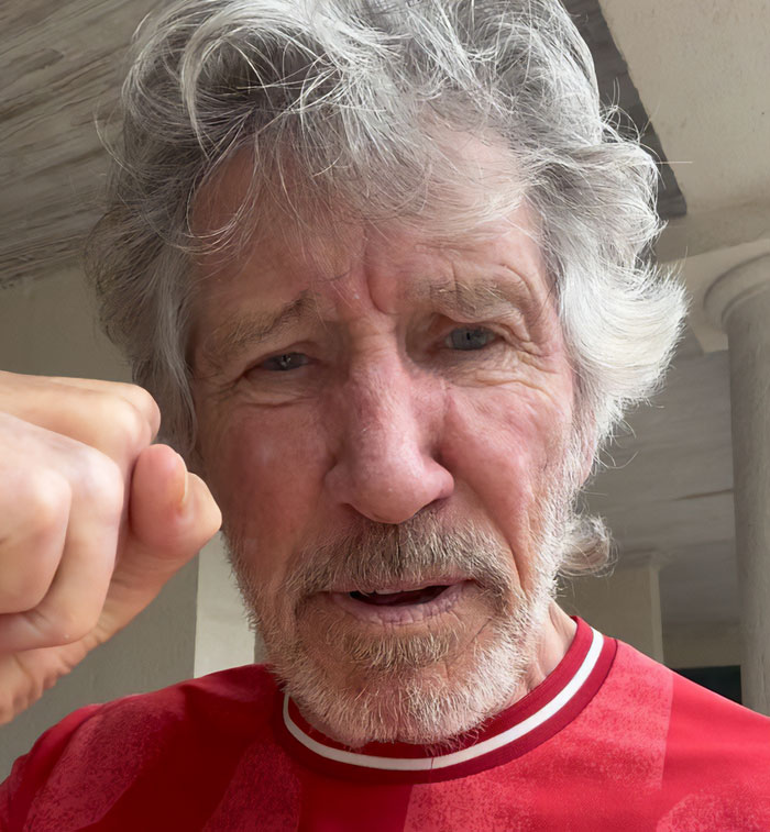 Roger Waters with a raised fist, expressing anger and intensity in a casual indoor setting.