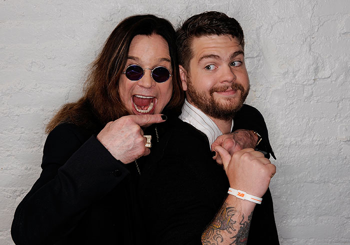 Ozzy Osbourne and his son posing playfully against a white brick wall, highlighting family and music legacy.