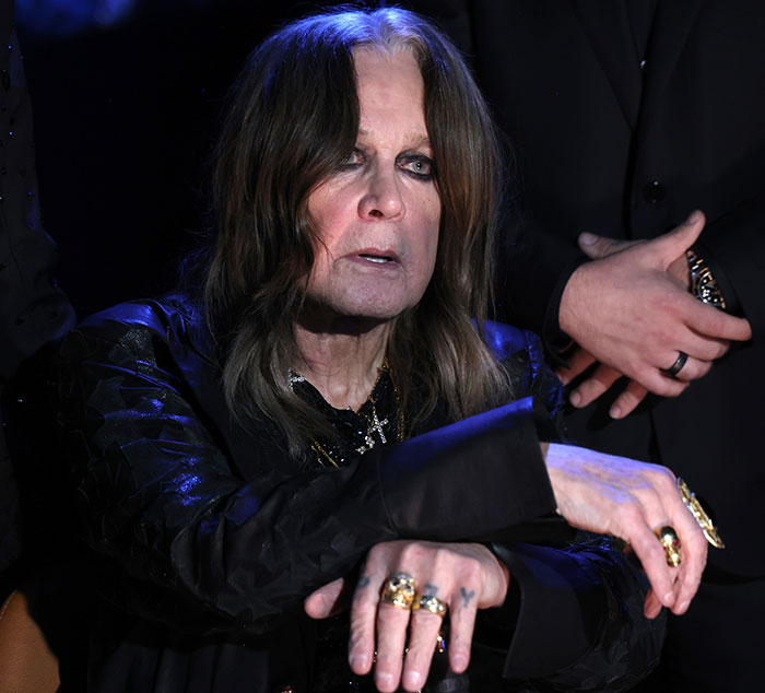 Ozzy Osbourne wearing rings and dark clothing with a serious expression amid a dramatic low-light setting.