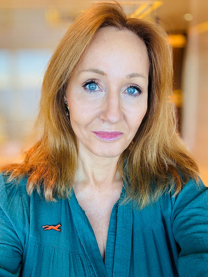 J.K. Rowling wearing teal top with red fox pin, facing camera with neutral expression in softly lit indoor setting. J.K. Rowling wearing teal top with red fox pin, facing camera with neutral expression in softly lit indoor setting.