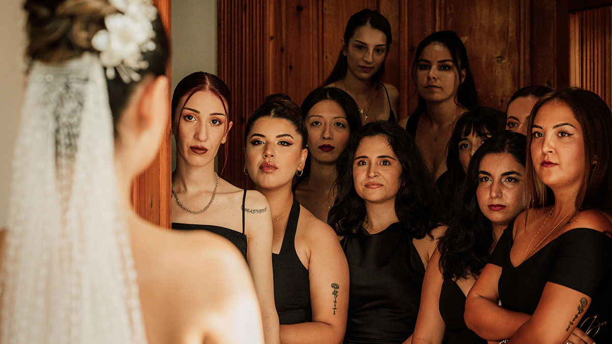 Bride in a white wedding dress with a pearl hairpiece facing a group of women dressed in black indoors.