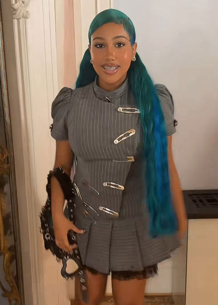 Young woman with long blue hair wearing a gray pinstripe dress with large safety pins, holding a black accessory indoors. Young woman with long blue hair wearing a gray pinstripe dress with large safety pins, holding a black accessory indoors.