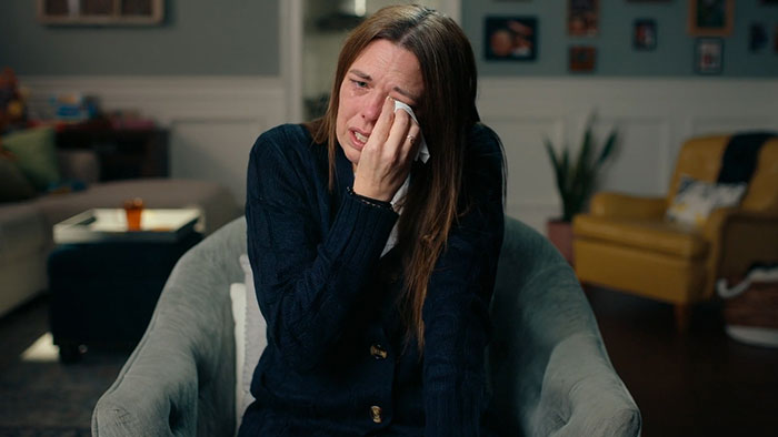 Mom who anonymously sent daughter lewd texts sitting in chair, tearfully wiping her face in a domestic setting. Mom who anonymously sent daughter lewd texts sitting in chair, tearfully wiping her face in a domestic setting.