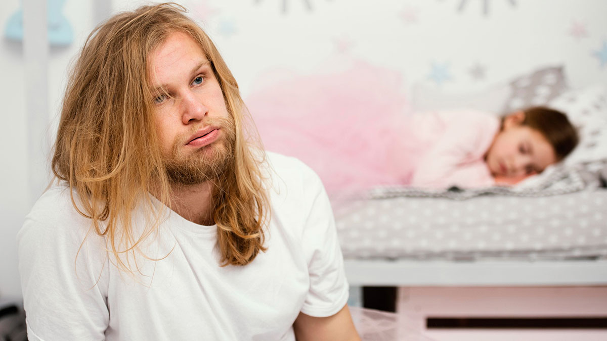 Man with long hair looking upset while a young girl sleeps in a bed, highlighting roommate conflict over sleeping arrangements
