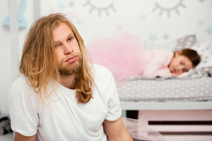 Young man sitting thoughtfully in a room while a child sleeps in a bed, reflecting tension over roommate sleeping arrangements. Young man sitting thoughtfully in a room while a child sleeps in a bed, reflecting tension over roommate sleeping arrangements.