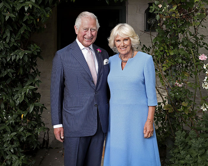 King Charles and Camilla standing together outdoors, smiling, with a garden background during a formal event. King Charles and Camilla standing together outdoors, smiling, with a garden background during a formal event.