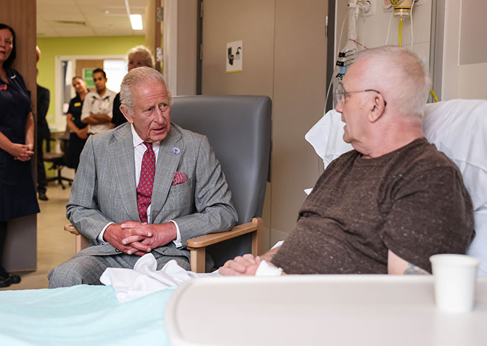 King Charles in a gray suit speaking with a patient in a hospital room, sharing an emotional cancer update. King Charles in a gray suit speaking with a patient in a hospital room, sharing an emotional cancer update.