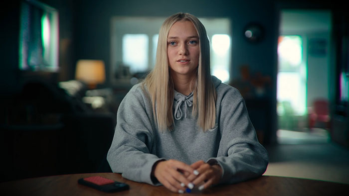 Young woman in a gray hoodie sitting at a table, sharing her story about a mom who sent lewd texts anonymously. Young woman in a gray hoodie sitting at a table, sharing her story about a mom who sent lewd texts anonymously.