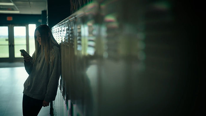 Teen girl leaning against school lockers, looking at phone, illustrating themes of secret torment and emotional distress. Teen girl leaning against school lockers, looking at phone, illustrating themes of secret torment and emotional distress.