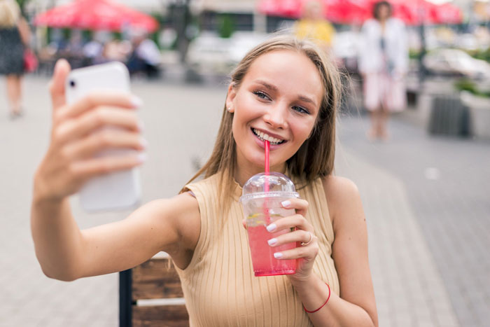 Young woman worried about online date behavior while taking selfie outdoors, holding a pink drink with a straw.