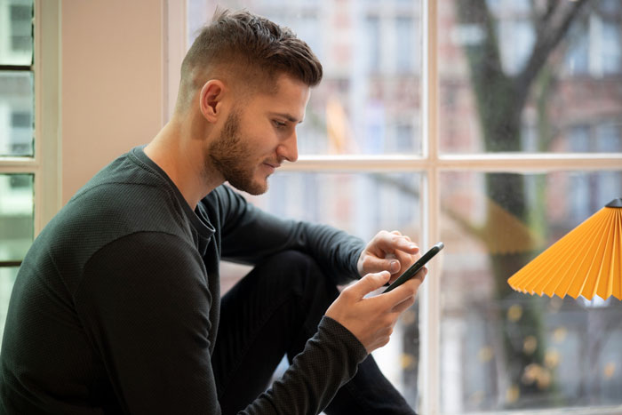 Young man sitting by a window, focused on his phone, illustrating lady worried over online date's behavior and memory of posts.