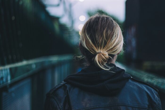 Person with a blonde hair tied back, wearing a black jacket, standing alone in an urban setting, reflecting on things never admitted.