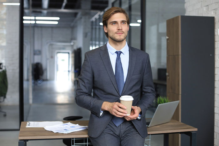 Young professional male dressed formally holding coffee cup in modern office, illustrating strict dress code compliance.