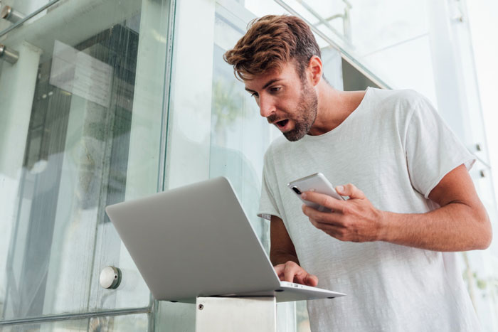 Man reacting with surprise at laptop while holding phone, illustrating employees maliciously complying with strict dress code.