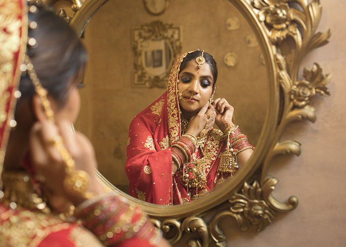 Woman in traditional bridal attire adjusting jewelry in front of an ornate mirror at a wedding event. Woman in traditional bridal attire adjusting jewelry in front of an ornate mirror at a wedding event.