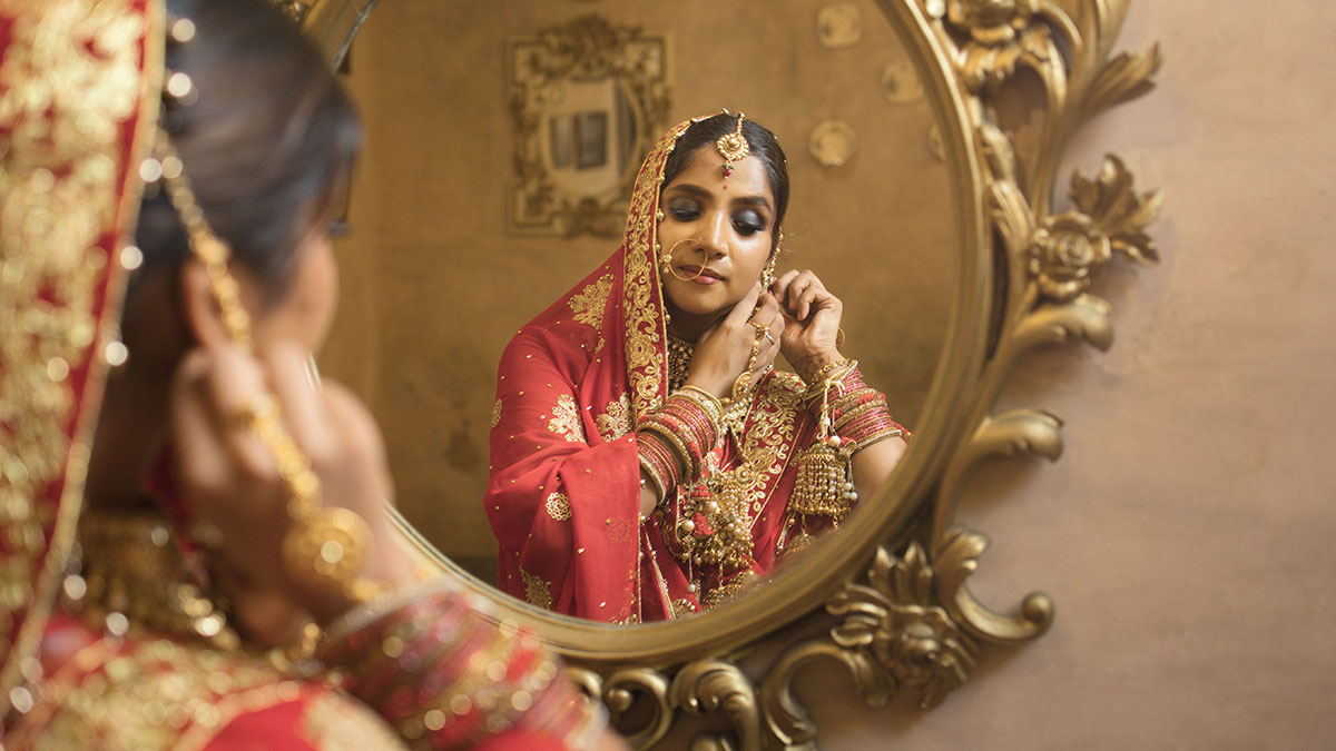 Woman dressed as bride adjusting jewelry in ornate mirror at sister-in-law wedding ceremony.