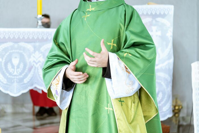 Priest wearing green liturgical vestments standing in a church interior with altar cloths and candles visible in background
