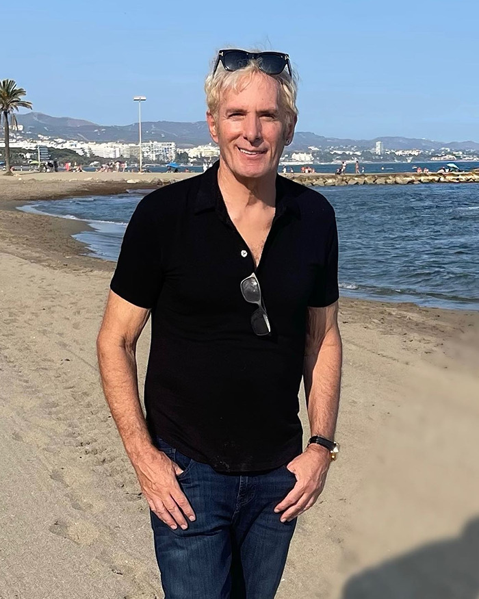 Michael Bolton standing on a beach wearing a black shirt and sunglasses with a scenic ocean background. Michael Bolton standing on a beach wearing a black shirt and sunglasses with a scenic ocean background.