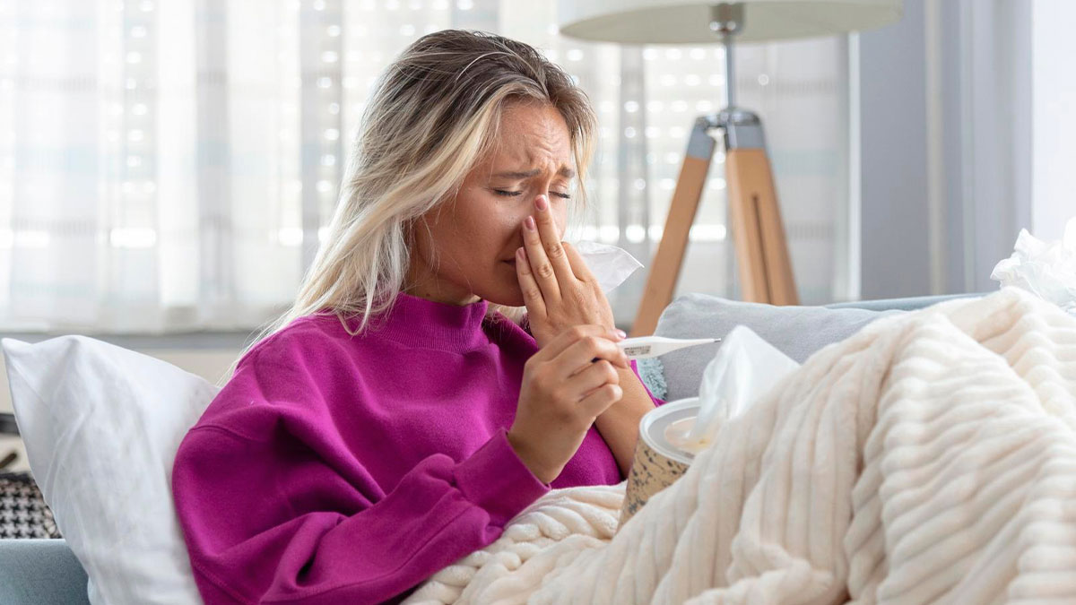 Woman with allergy symptoms on couch, holding tissue and thermometer, showing distress over allergy-safe food sabotage.
