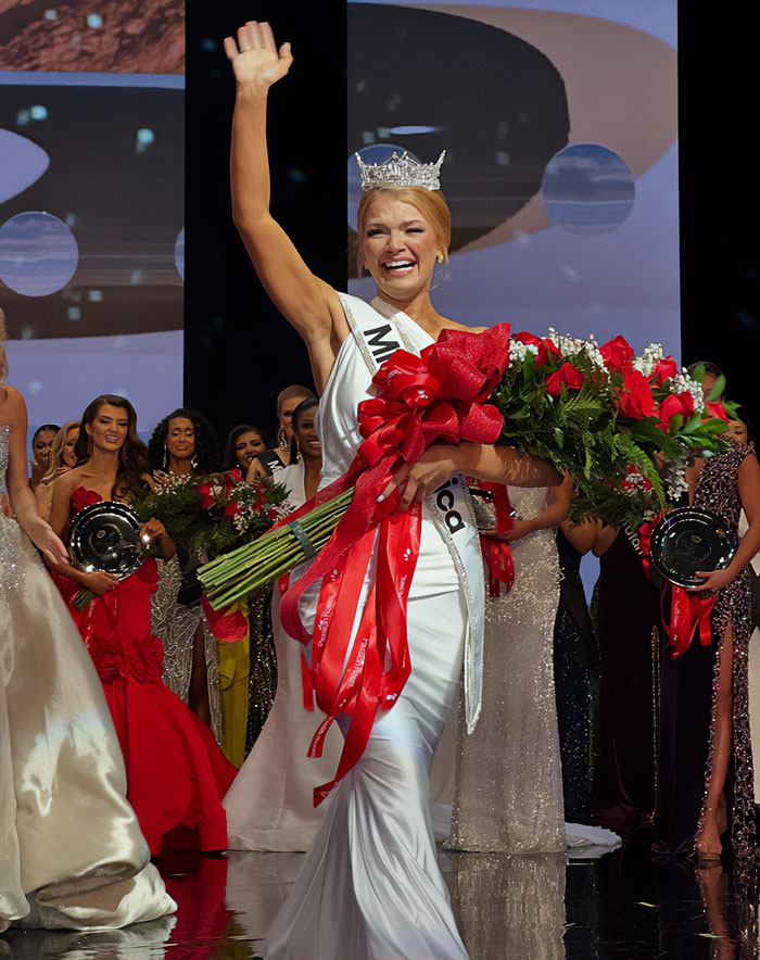 Miss America winner wearing crown and sash, holding bouquet, waving to crowd amid pageant participants on stage. Miss America winner wearing crown and sash, holding bouquet, waving to crowd amid pageant participants on stage.