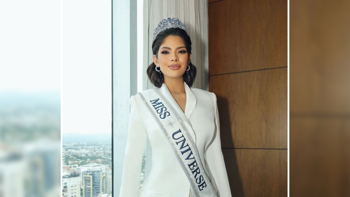 Miss Universe winner wearing crown and sash in a white suit, posing confidently indoors with city view in background