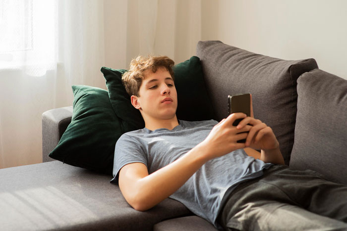 Teenage son lying on couch using phone, showing refusal for family bonding and emotional distance from mother. Teenage son lying on couch using phone, showing refusal for family bonding and emotional distance from mother.