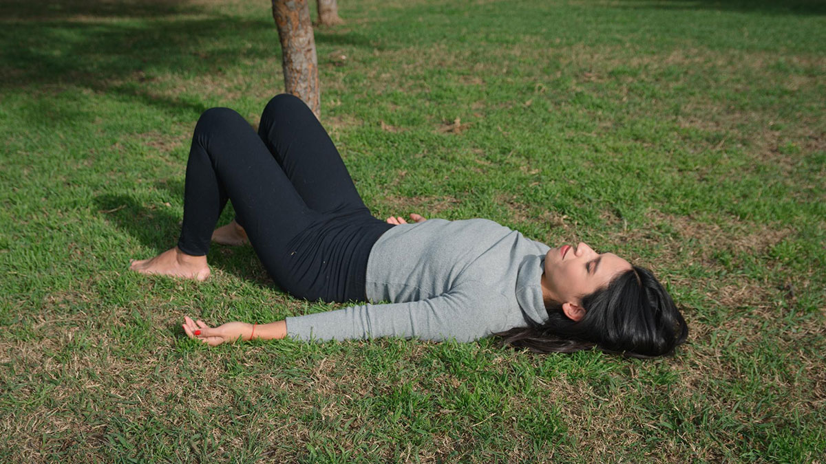 Teen lying on grass after fainting and hurting herself, in distress near a tree outdoors on a sunny day.
