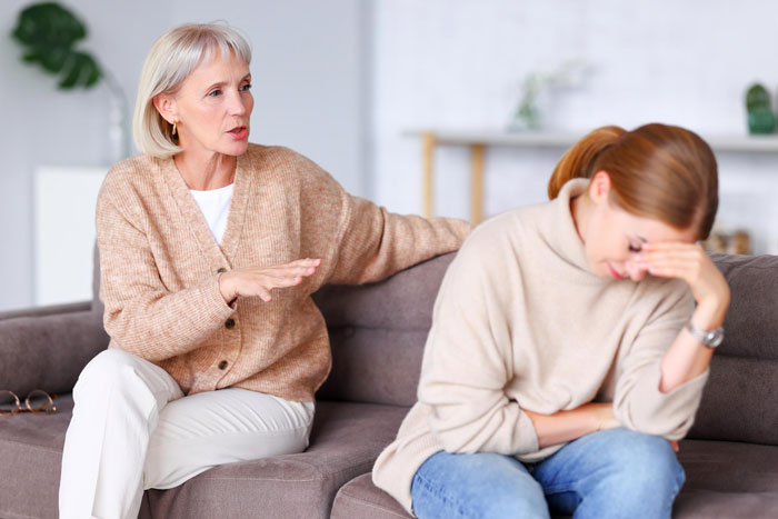 Teen sitting upset on couch holding head as older woman speaks and comforts her in a living room setting.