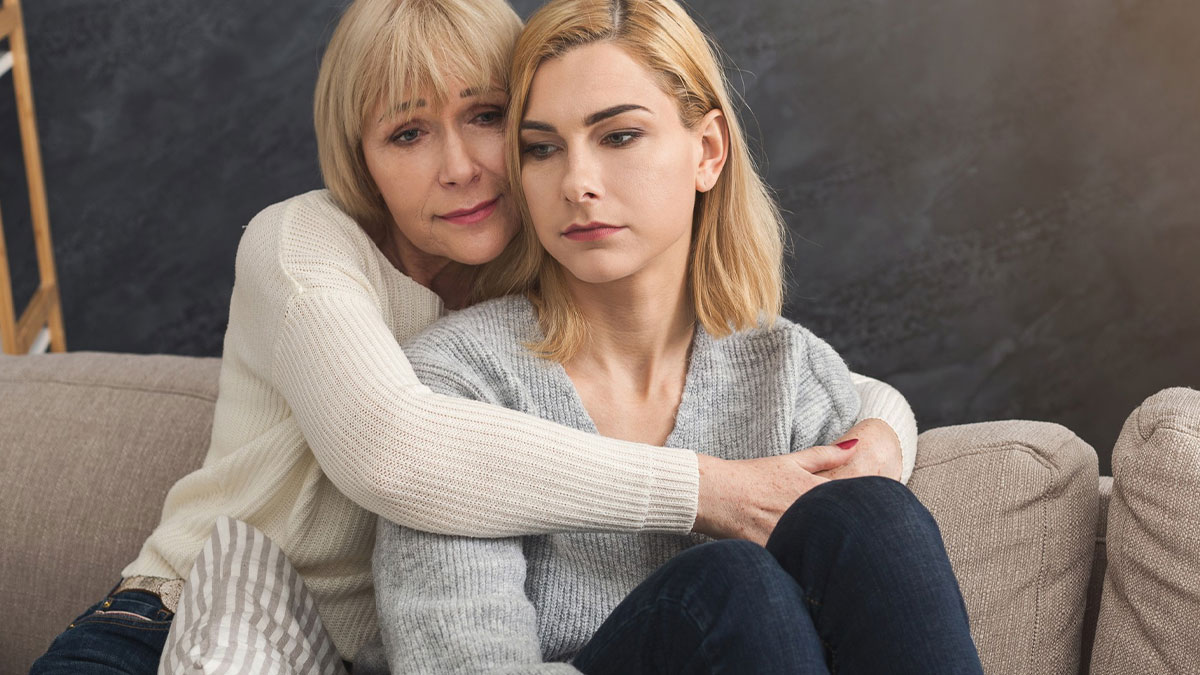 Older woman hugging her adult daughter on a couch, showing support and care in a mom retirement plan moment.