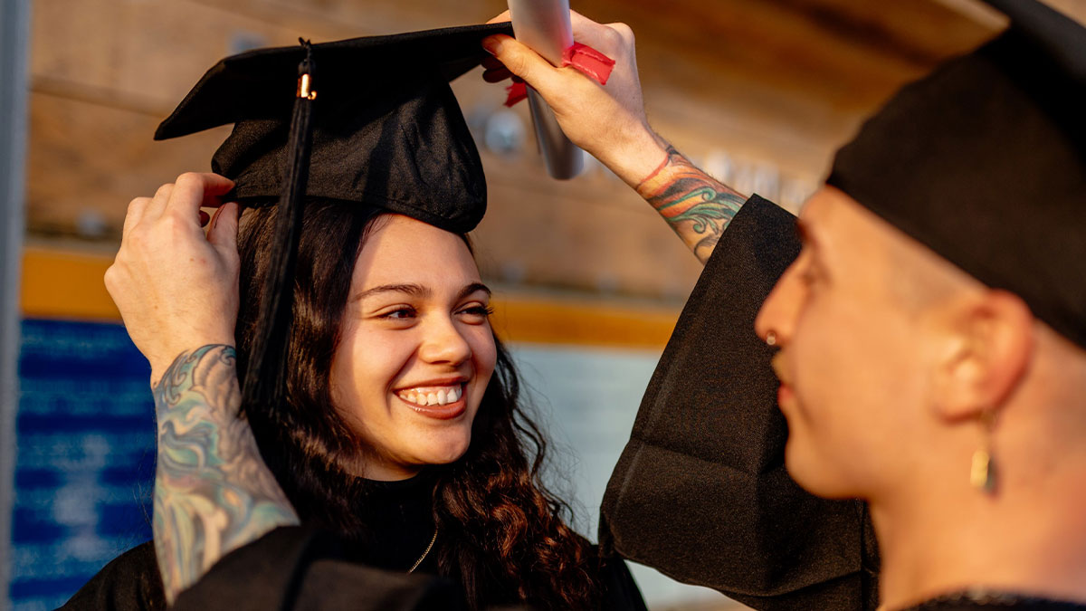 Two graduates in caps and gowns smiling as one adjusts the other's graduation cap in an outdoor setting.