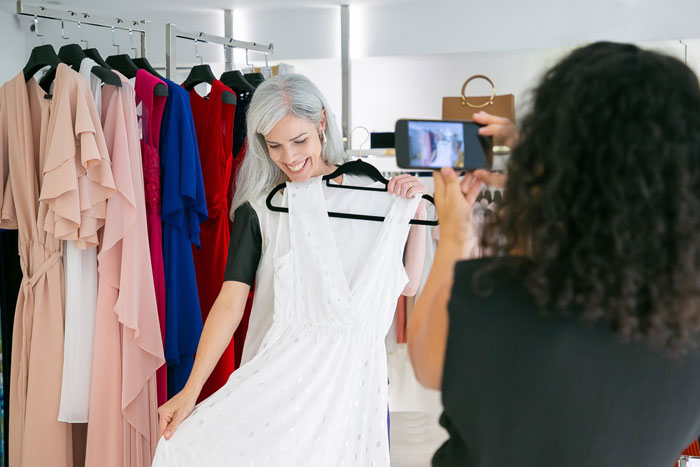 Woman holding a white dress in a boutique while another person takes a photo, related to rude MIL regrets wearing white to wedding. Woman holding a white dress in a boutique while another person takes a photo, related to rude MIL regrets wearing white to wedding.