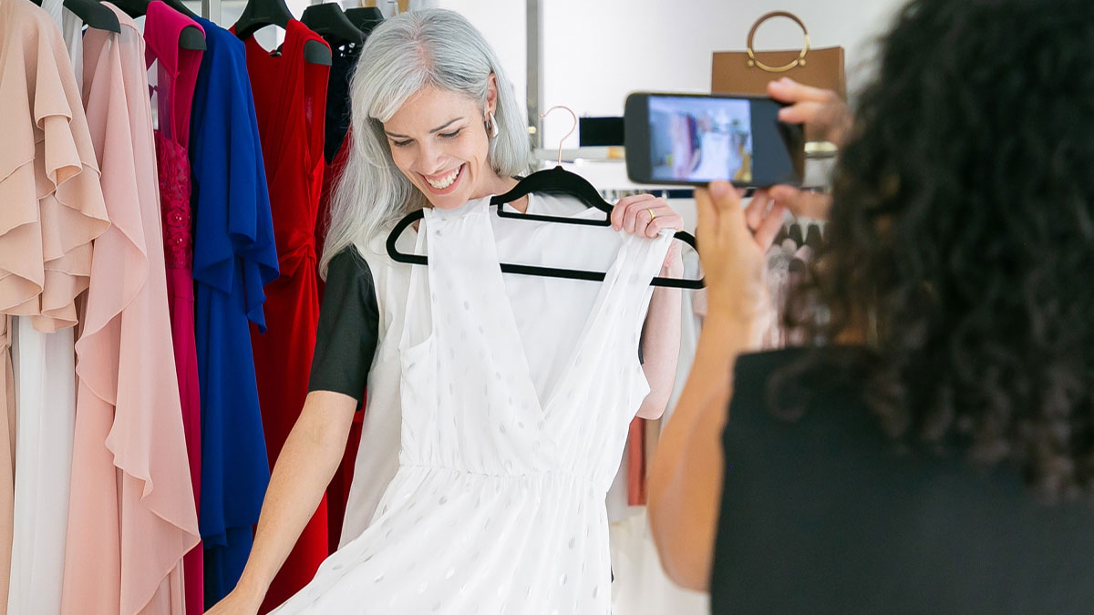 Woman holding a white dress in a boutique while another person takes a photo, highlighting bridal party and wedding dress issues.