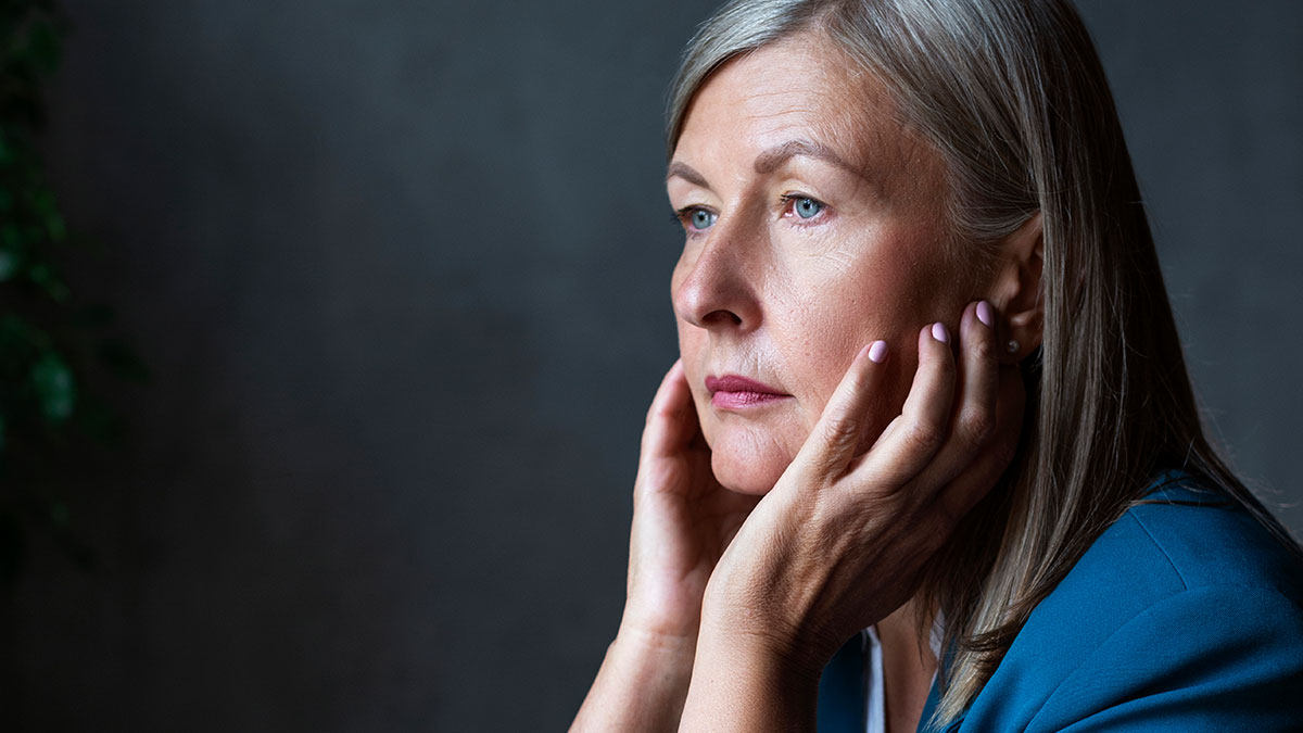 Woman with gray hair in a blue jacket looking pensive, holding her face with both hands in a dark background, mother poisoned me.