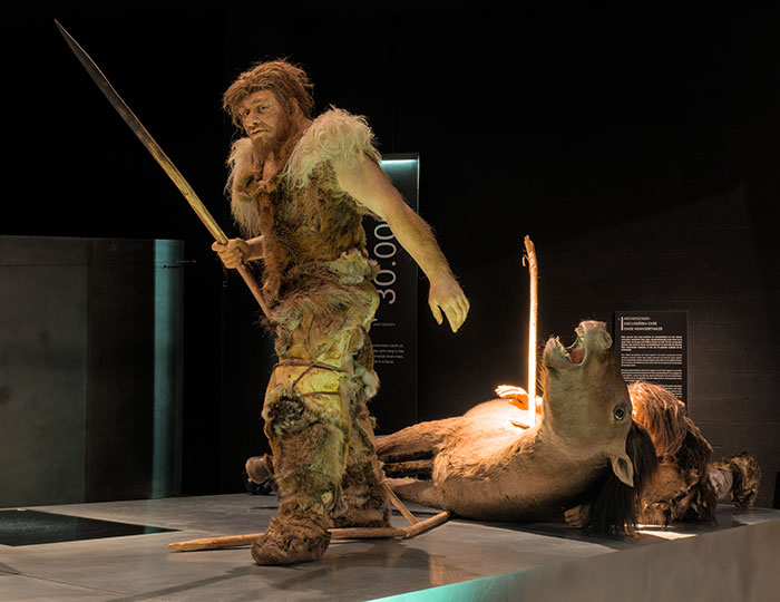 Reconstructed Petralona Man figure holding spear beside prehistoric animal in a dimly lit museum exhibit. Reconstructed Petralona Man figure holding spear beside prehistoric animal in a dimly lit museum exhibit.