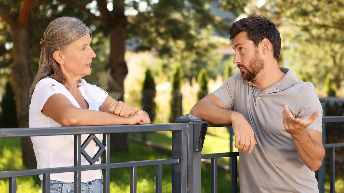 Neighbor upset talking to man over nanny parking dispute in front of home near metal fence outdoors.