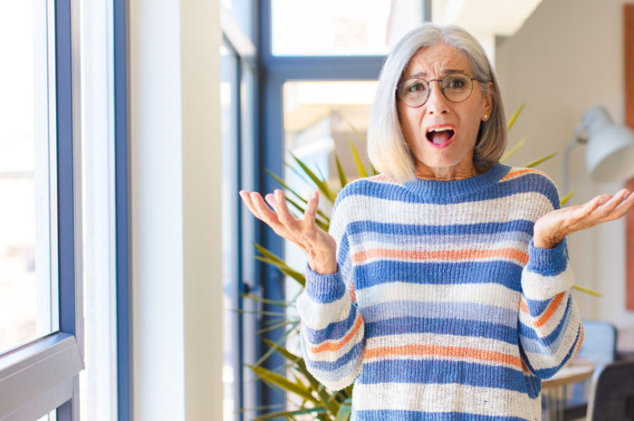 Older woman in striped sweater and glasses looking distressed indoors, illustrating nosy neighbor trauma concept. Older woman in striped sweater and glasses looking distressed indoors, illustrating nosy neighbor trauma concept.