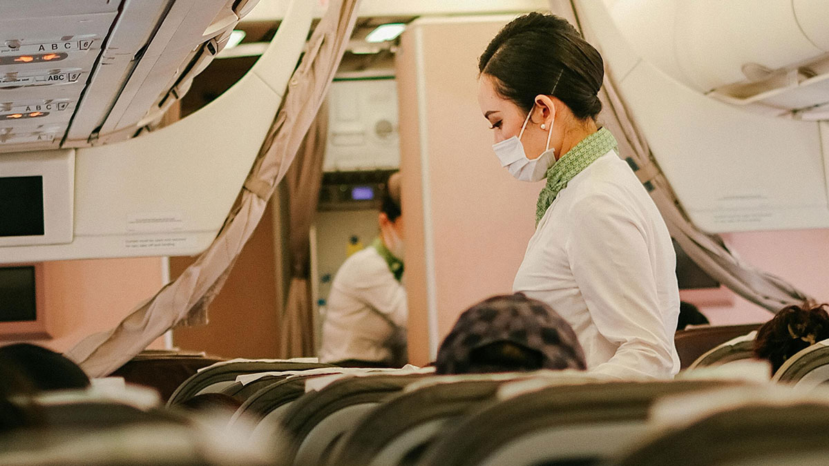 Flight attendant wearing mask assisting passengers inside an airplane cabin during boarding process.