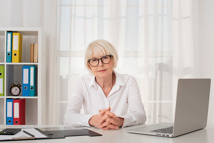 Older female teacher in glasses sitting at desk with laptop, representing a classroom incident involving a student and suspension issue. Older female teacher in glasses sitting at desk with laptop, representing a classroom incident involving a student and suspension issue.