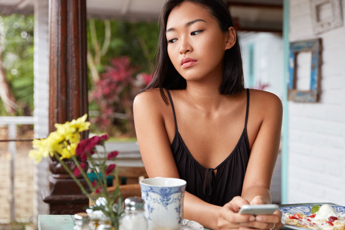 Young woman holding phone at a table with food, appearing thoughtful while considering ordering fried pork intestine. Young woman holding phone at a table with food, appearing thoughtful while considering ordering fried pork intestine.