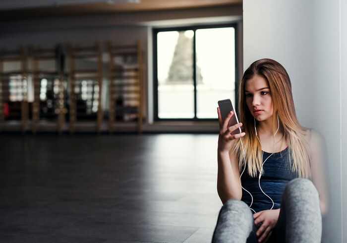 Young woman sitting alone in a gym, looking at her phone, illustrating Gen Z clients troubling behavior and mental health. Young woman sitting alone in a gym, looking at her phone, illustrating Gen Z clients troubling behavior and mental health.