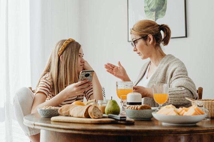 A 19-year-old girl looks irked while talking to a woman over breakfast, reflecting forced parenting of little sister. A 19-year-old girl looks irked while talking to a woman over breakfast, reflecting forced parenting of little sister.