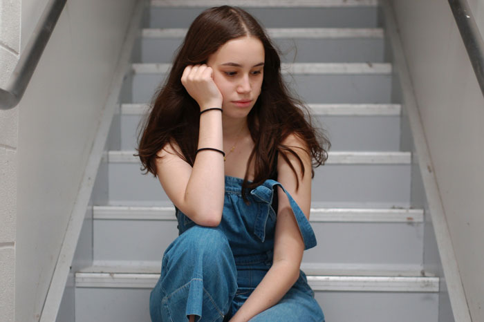 Teenage girl in denim sitting on stairs, looking upset about being forced to be a parent to her little sister. Teenage girl in denim sitting on stairs, looking upset about being forced to be a parent to her little sister.