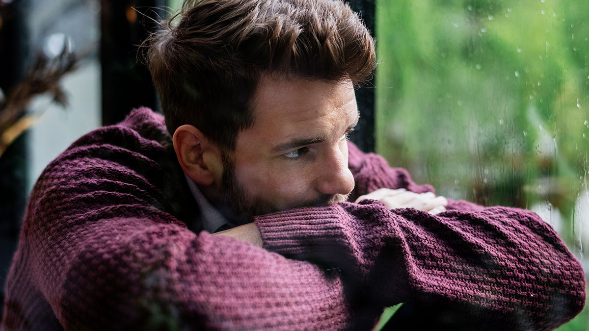 Young man in a maroon sweater looking out a rainy window, reflecting on caring for his autistic brother and family pressure.