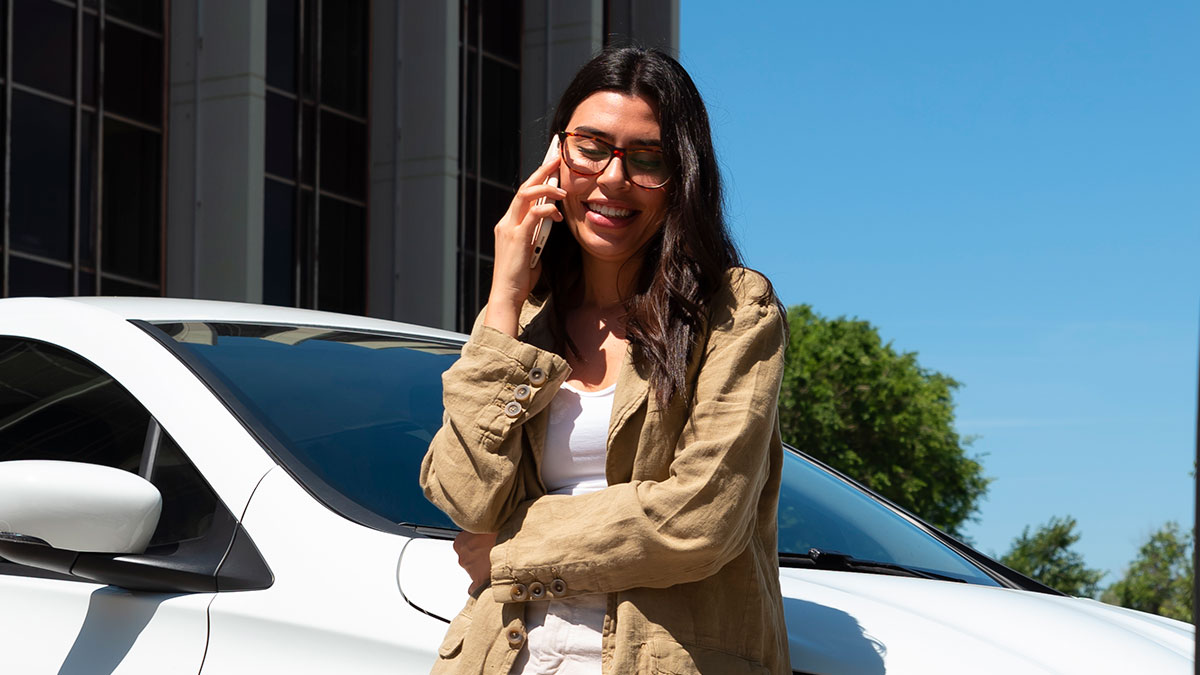 Woman with glasses smiling and talking on phone next to white car parked in EV charging spot outdoors.
