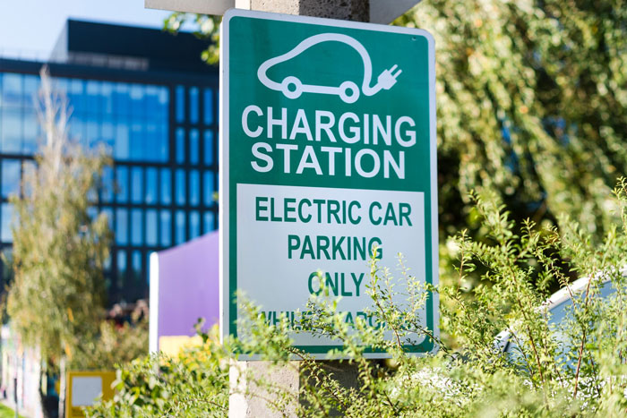 Green electric car charging station sign indicating parking for EVs only near a building with trees and bushes around. Green electric car charging station sign indicating parking for EVs only near a building with trees and bushes around.