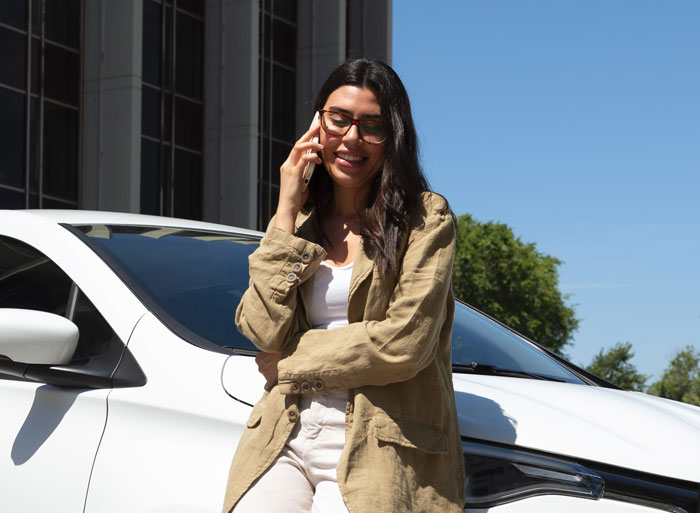 Woman talking on phone leaning against white car parked in EV charging spot on a sunny day outdoors. Woman talking on phone leaning against white car parked in EV charging spot on a sunny day outdoors.