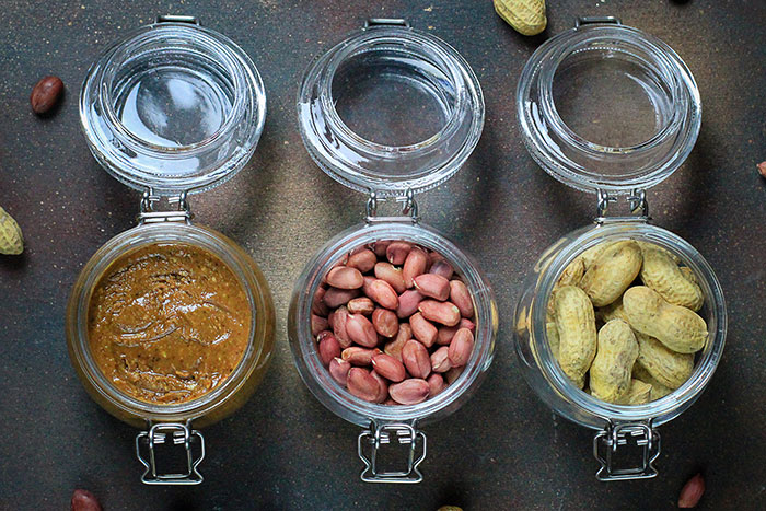 Three glass jars containing peanut butter, shelled peanuts, and unshelled peanuts on a dark surface, highlighting peanut allergy risk.