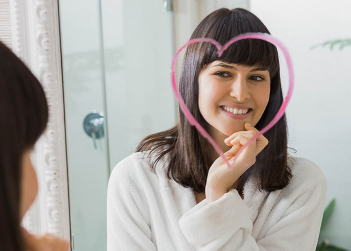 Woman smiling in a bathroom mirror with a pink heart drawn on the glass representing life hacks that actually work.
