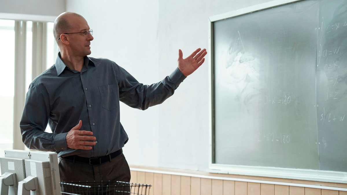 Teacher in a classroom pointing at a chalkboard sharing funny jokes and iconic comments to surprise students.